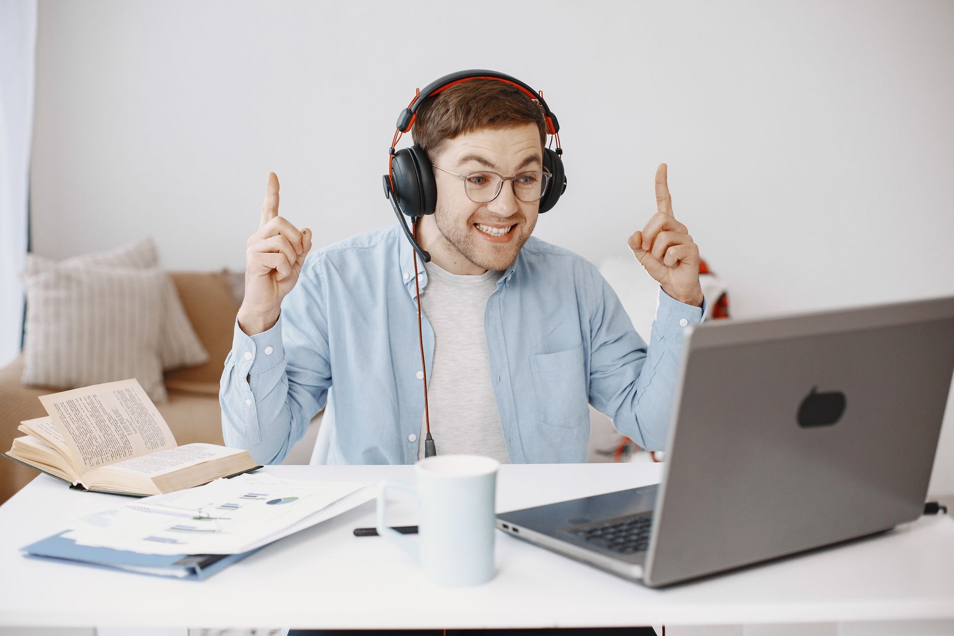 Smiling young man with headphones working remotely with a laptop and book on desk.