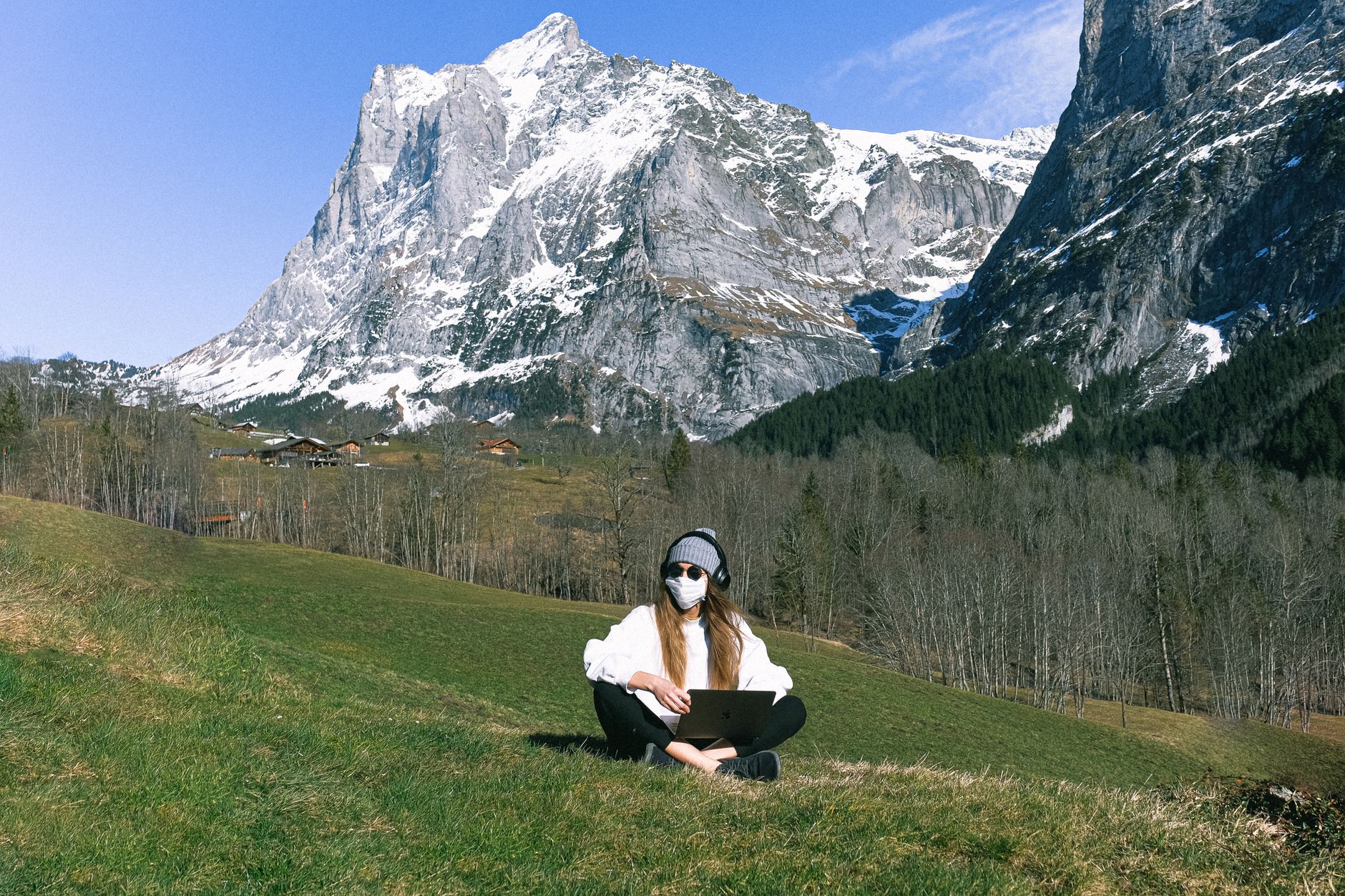 A woman sits with a laptop wearing a mask in a scenic mountain landscape, embracing remote work outdoors.