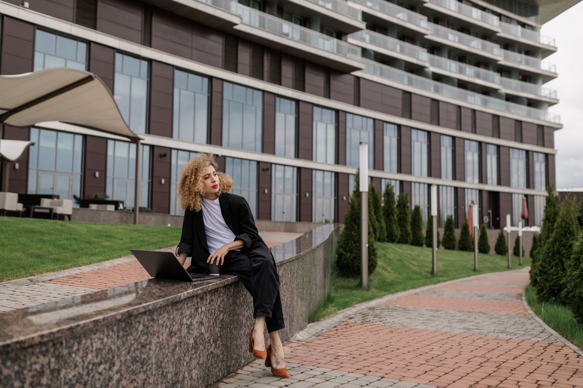 Businesswoman using a laptop outside a modern building, wearing black blazer and stylish heels.