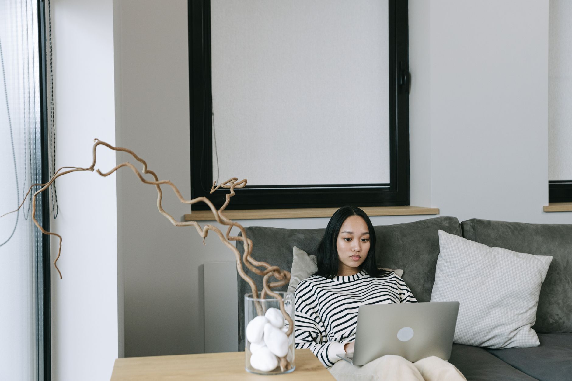 Asian woman working from home on laptop, sitting on a sofa in a modern, minimalist space.