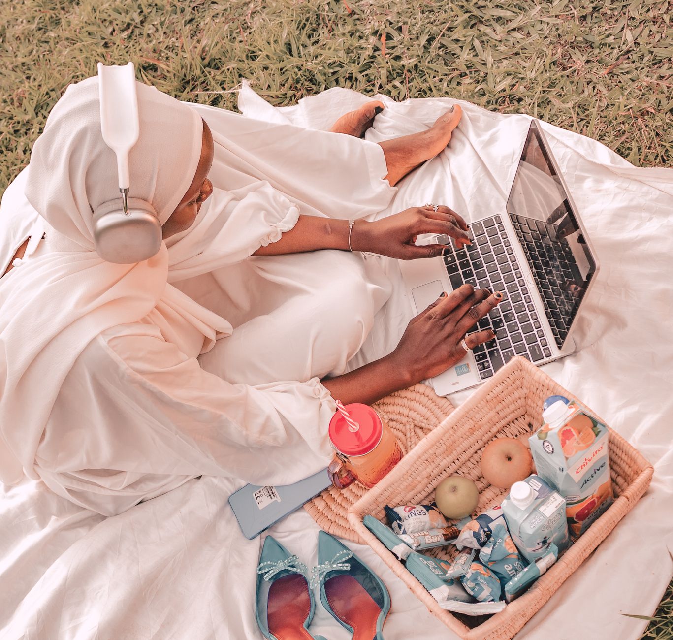 African woman using a laptop during a picnic in a sunny park, enjoying snacks and technology.