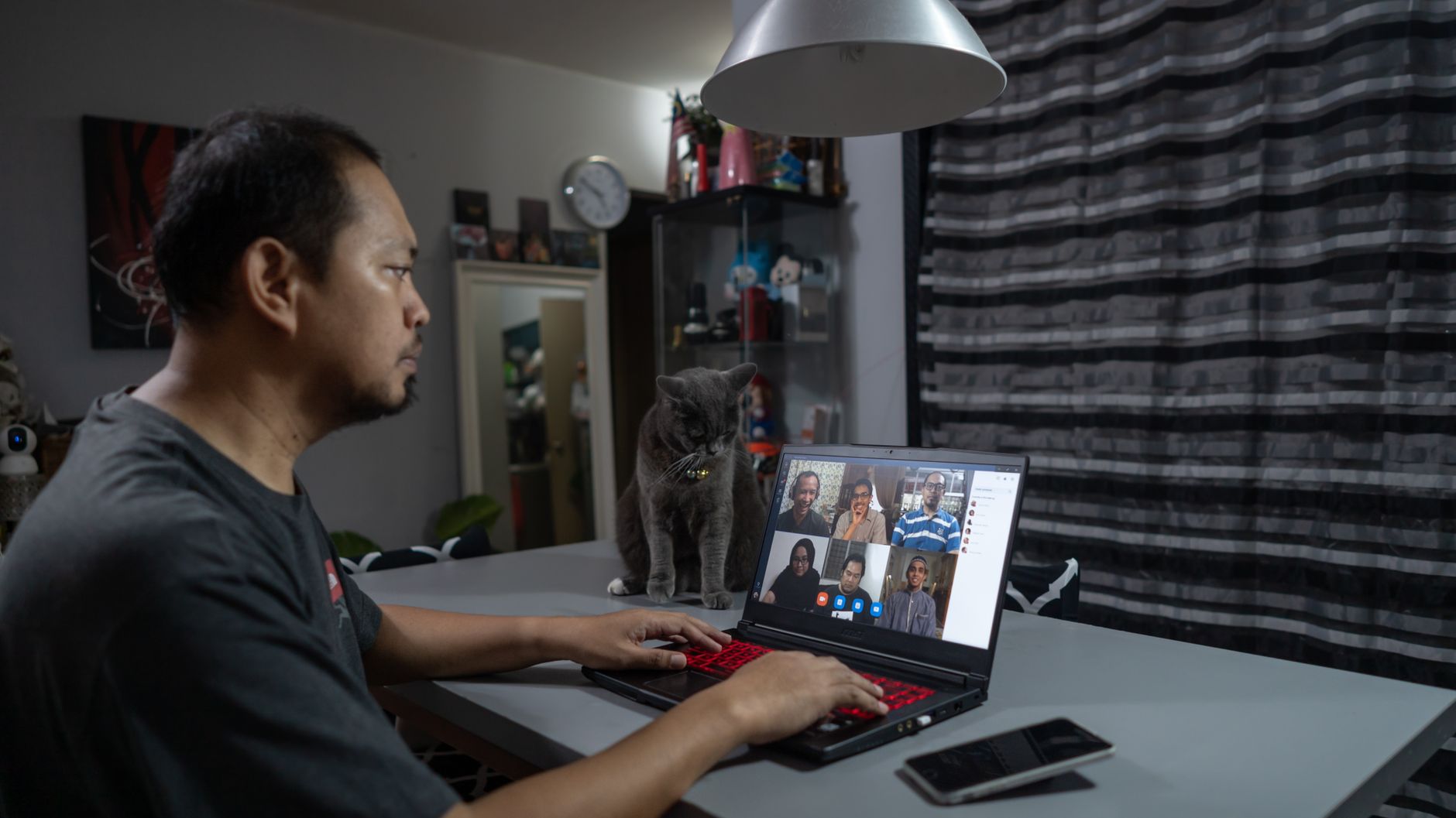 Asian man working remotely on a laptop video call from home with a cat nearby.