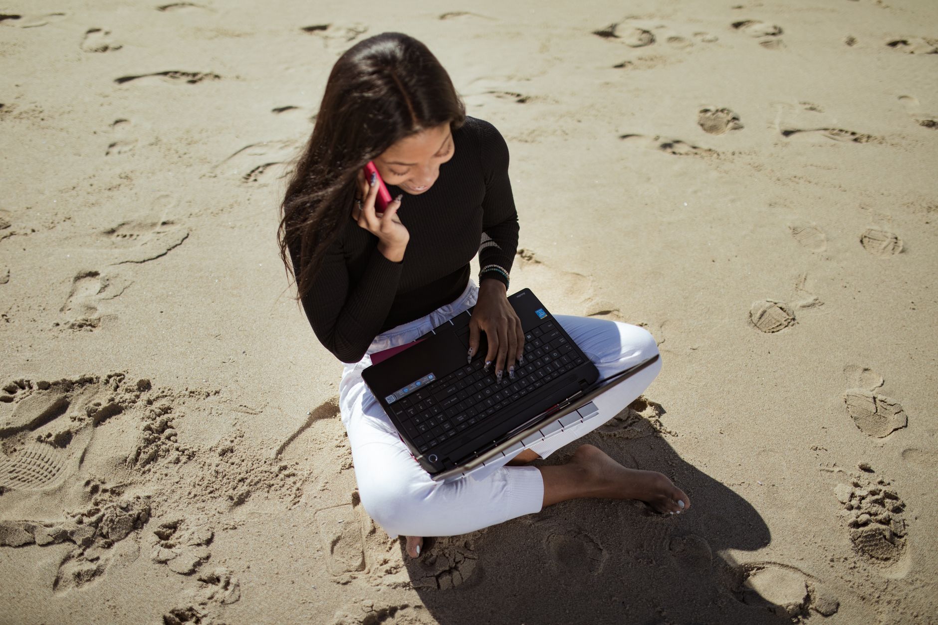 Woman working remotely on a beach, using a laptop and talking on the phone.