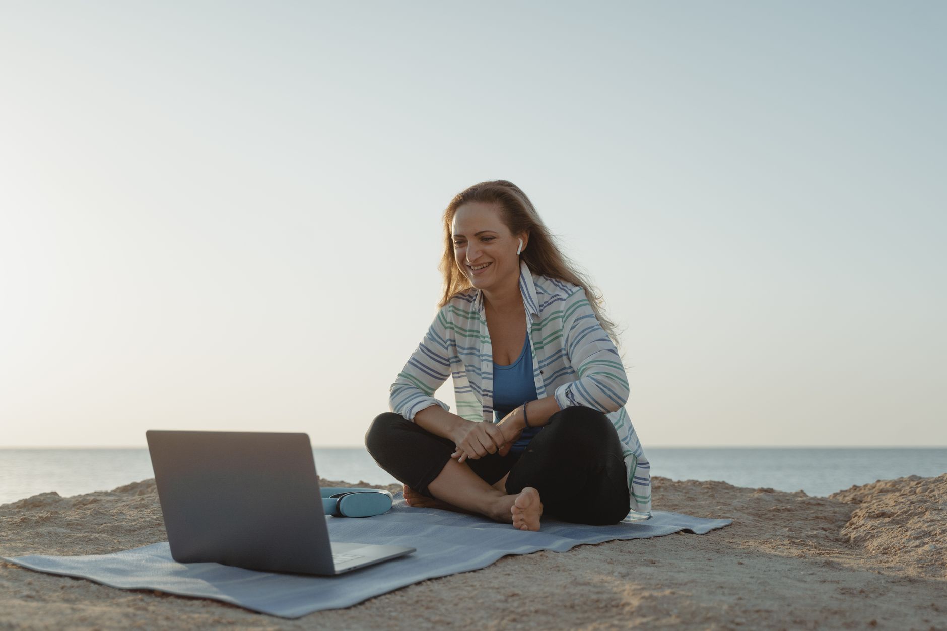 Woman sitting on a beach using a laptop while wearing wireless earbuds, enjoying a relaxing work setting.