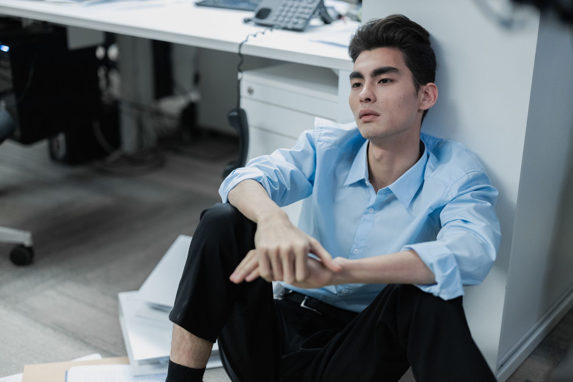 An exhausted Asian man sits on an office floor looking reflective and stressed, surrounded by paperwork.
