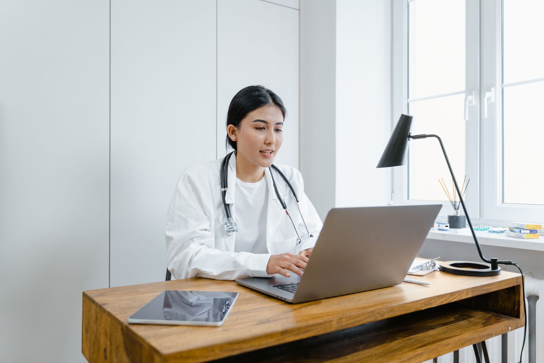 A female doctor in a white coat uses a laptop for an online consultation from her office.