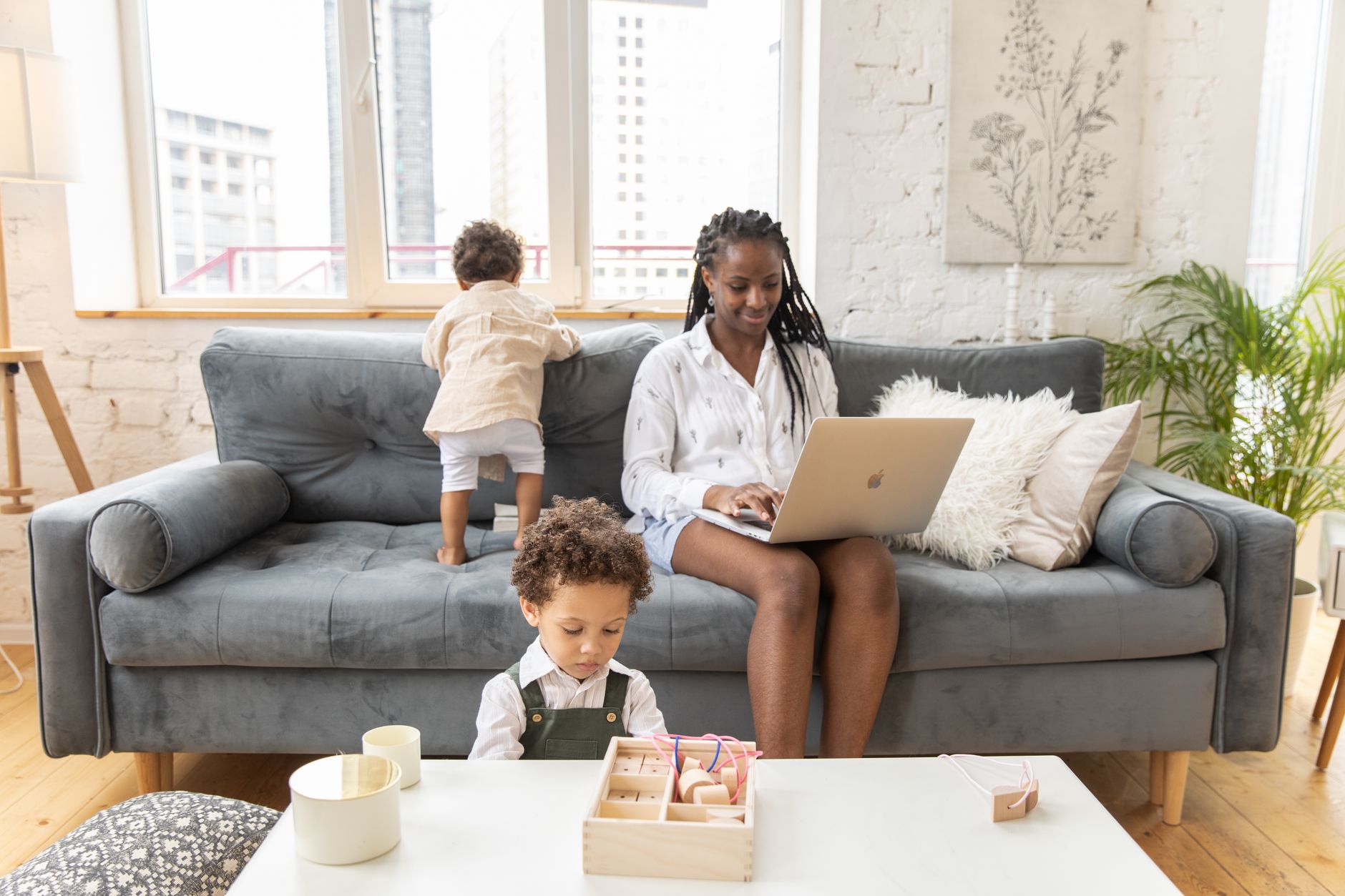 A mother works via laptop while her young children play in the living room.