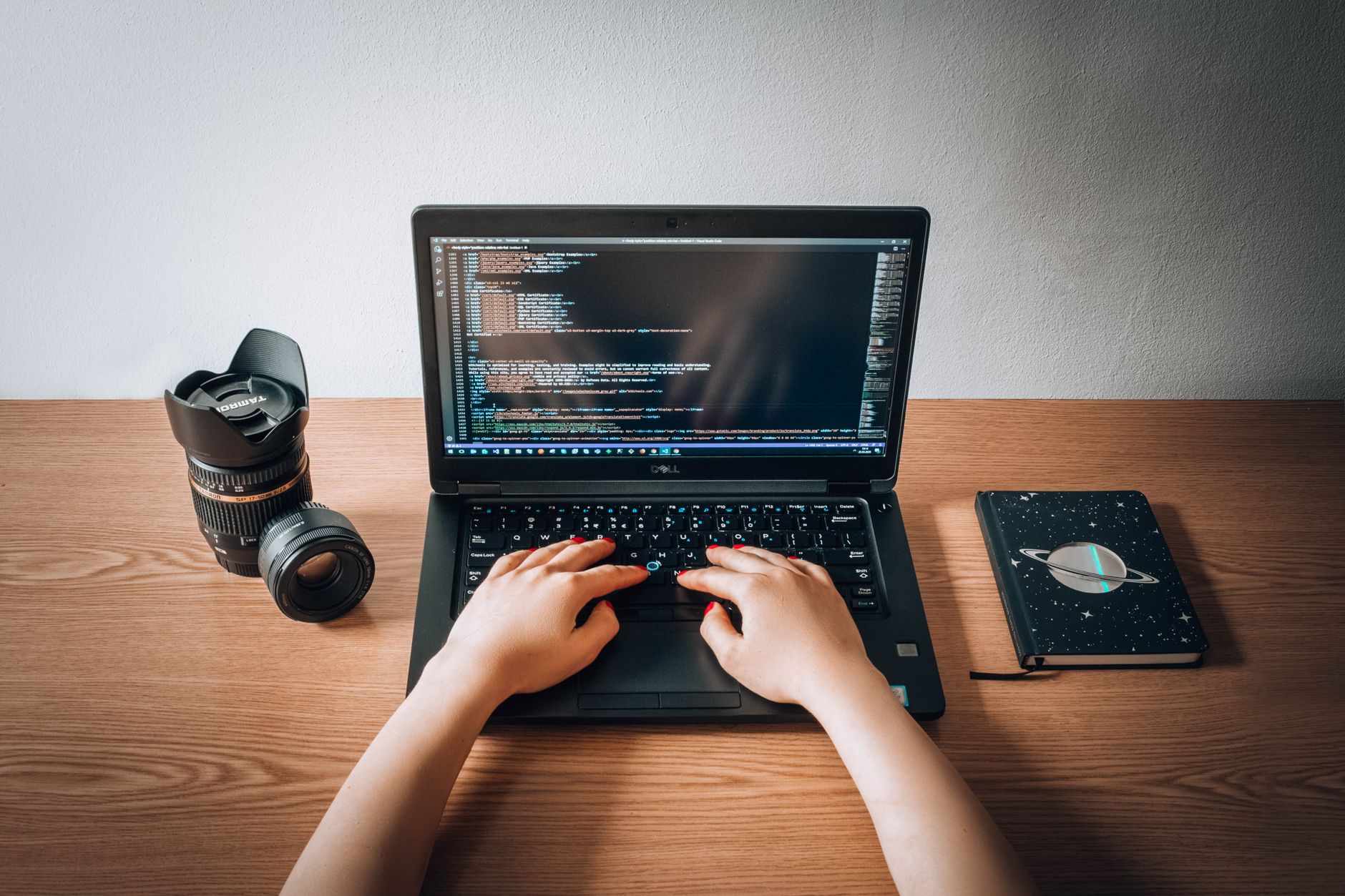 A woman coding on a laptop with a camera lens and notebook on a wooden desk in a home office.