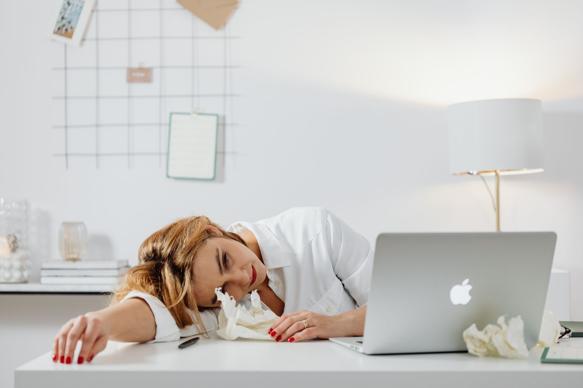 Exhausted woman in white attire rests head on desk beside laptop and crumpled papers.