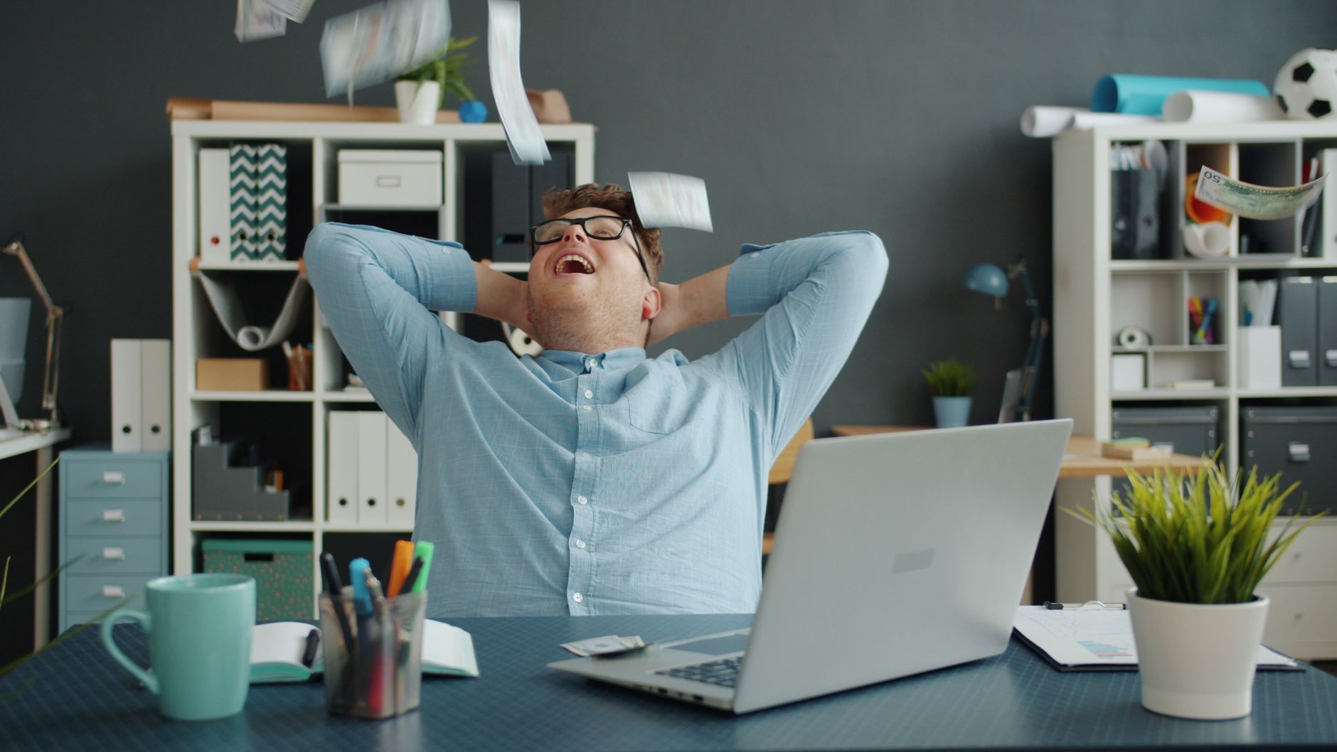 A happy businessman relaxes at his desk as banknotes fall around him, symbolizing success.
