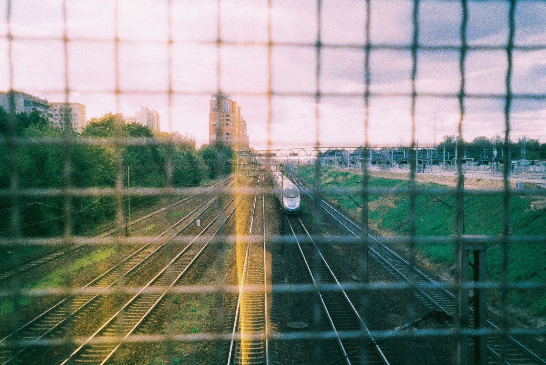 A high-speed train travels through Warsaw, Poland at sunset, captured from behind a fence.
