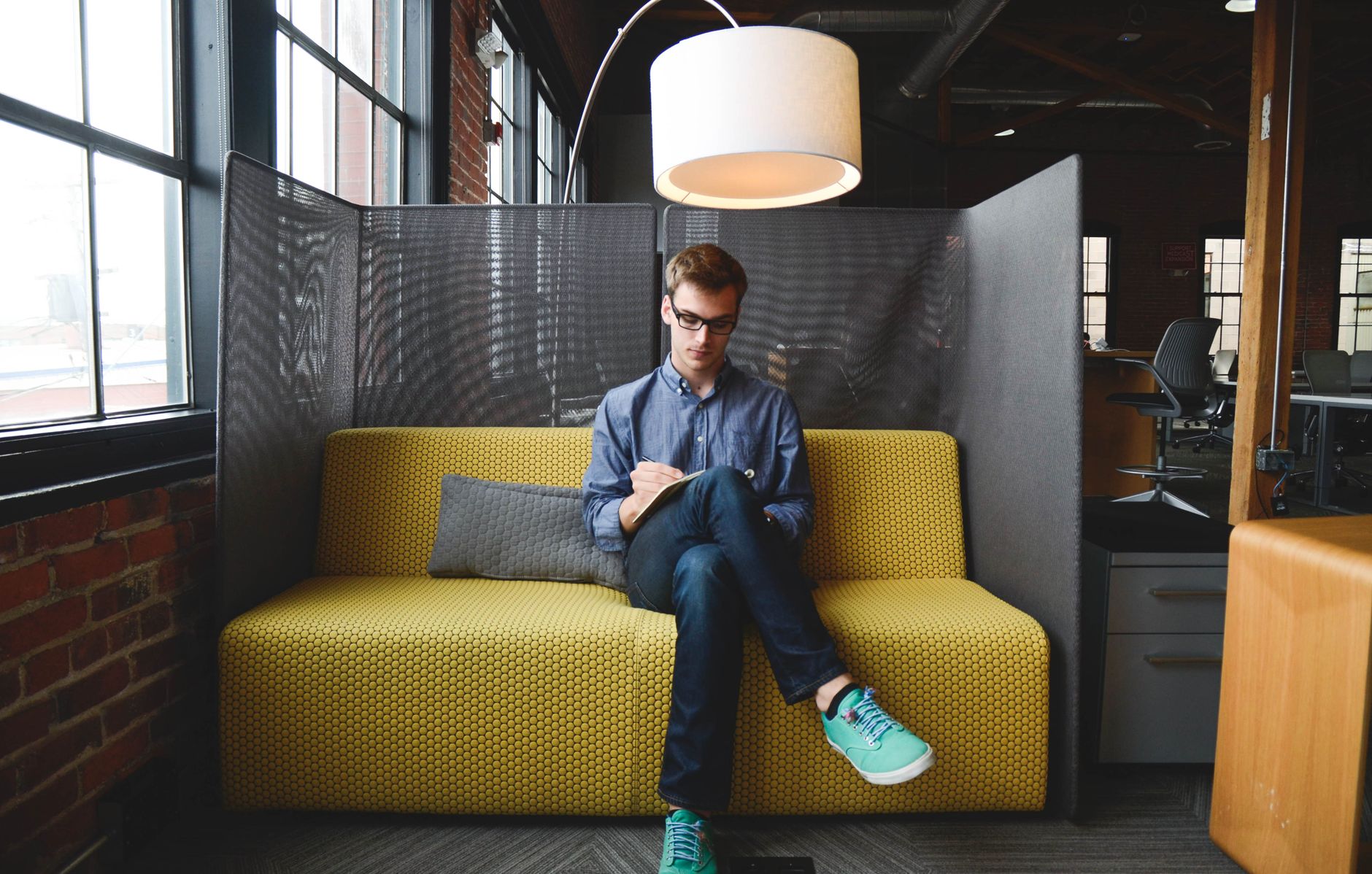 Young man writing on a notebook while sitting on a couch in a modern coworking space.