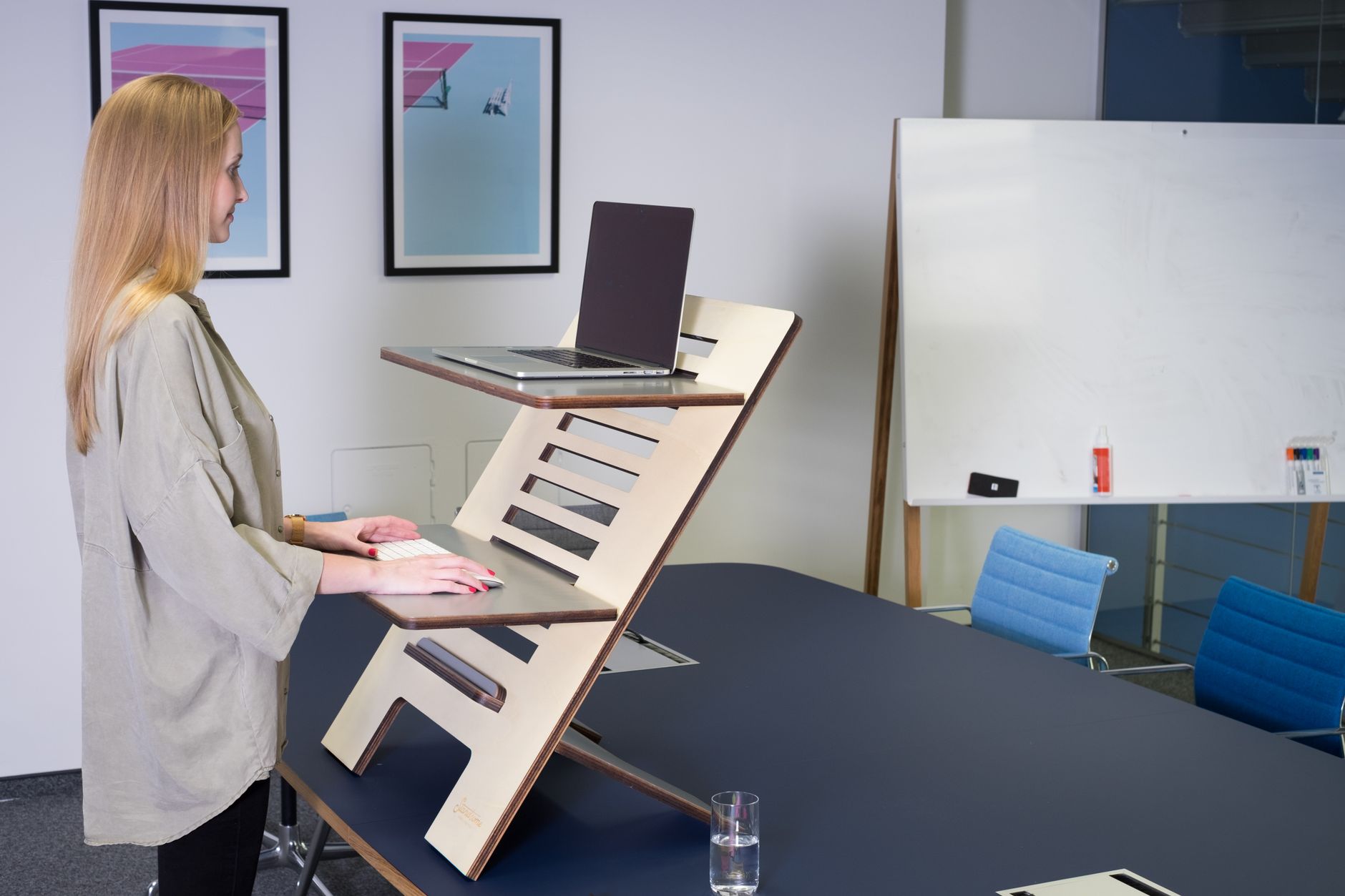 Woman using a standing desk with laptop in a modern office setting, promoting ergonomic workspace solutions.