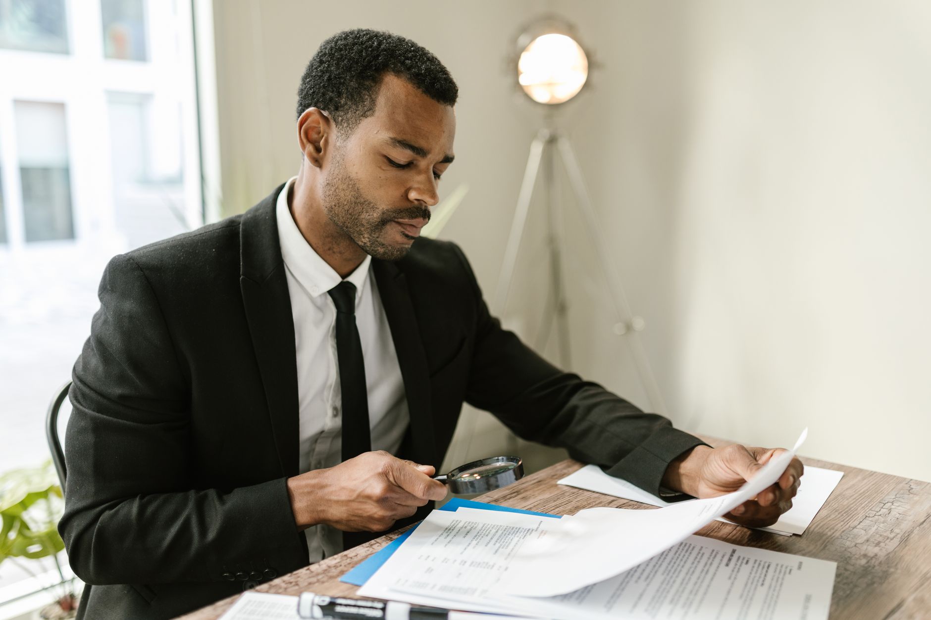 Professional man examines paperwork with magnifying glass at office desk, highlighting attention to detail.