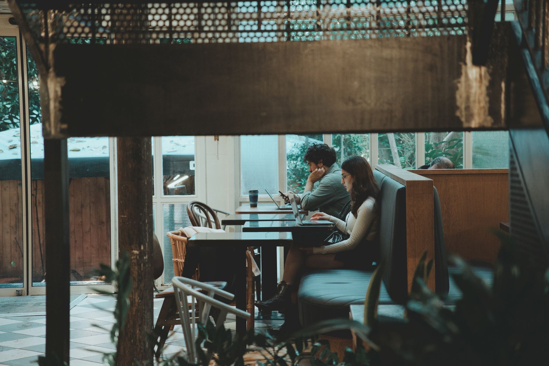 Two young adults working on laptops in a stylish, cozy café interior surrounded by plants.