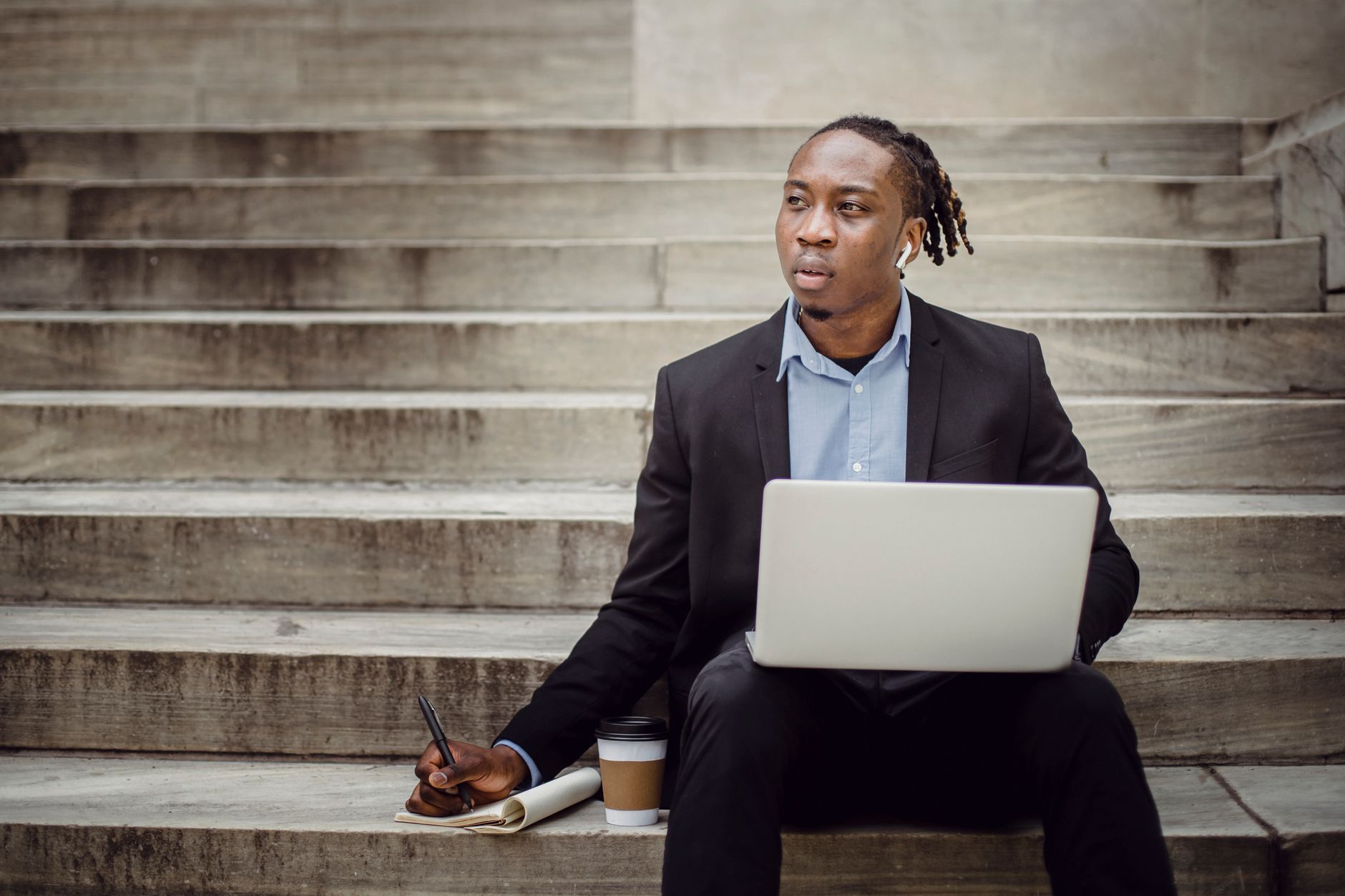 Concentrated young ethnic male freelancer thinking about business strategy and taking notes in planner while working on laptop sitting on st