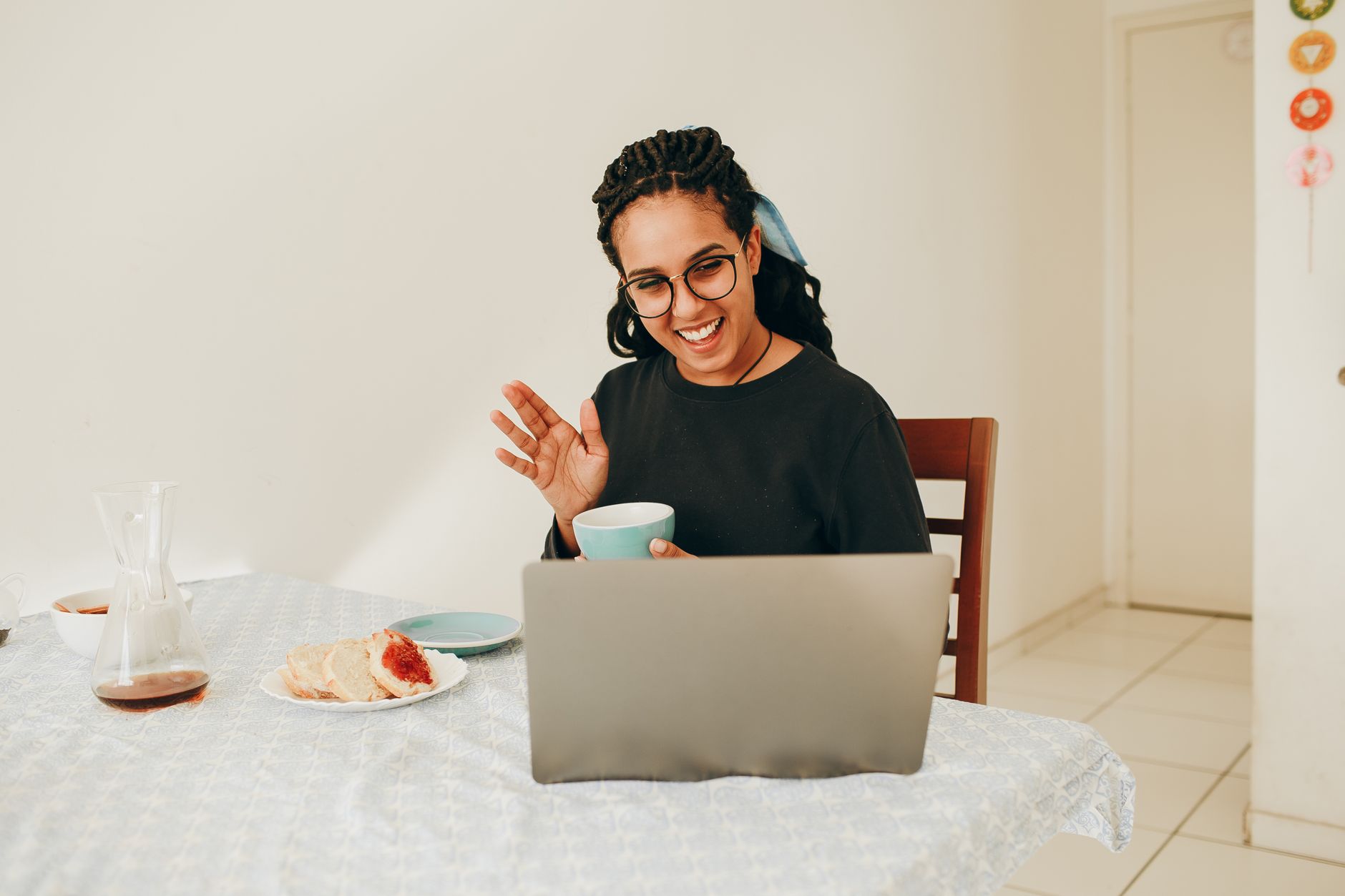 A woman enjoys a virtual coffee meeting in a cozy home setting. Smiling and waving at the laptop.
