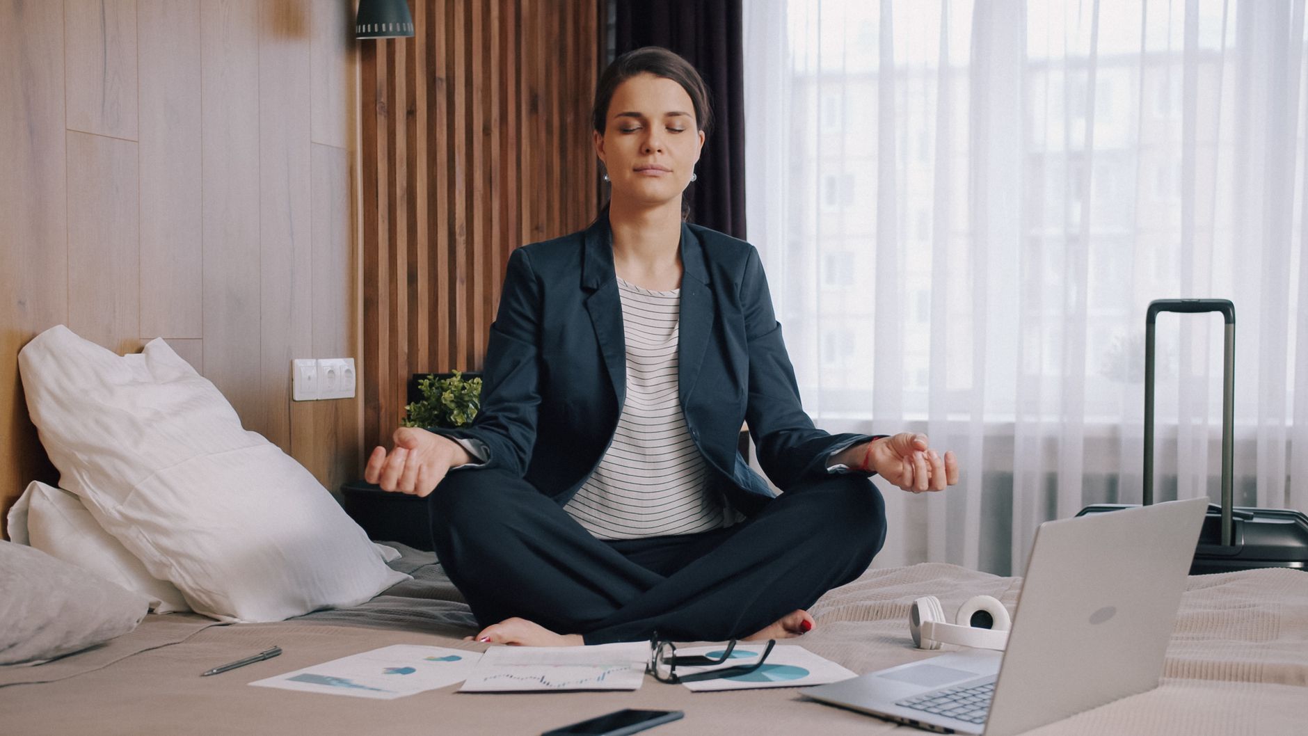 Businesswoman practicing meditation on bed in hotel room, achieving work-life balance.
