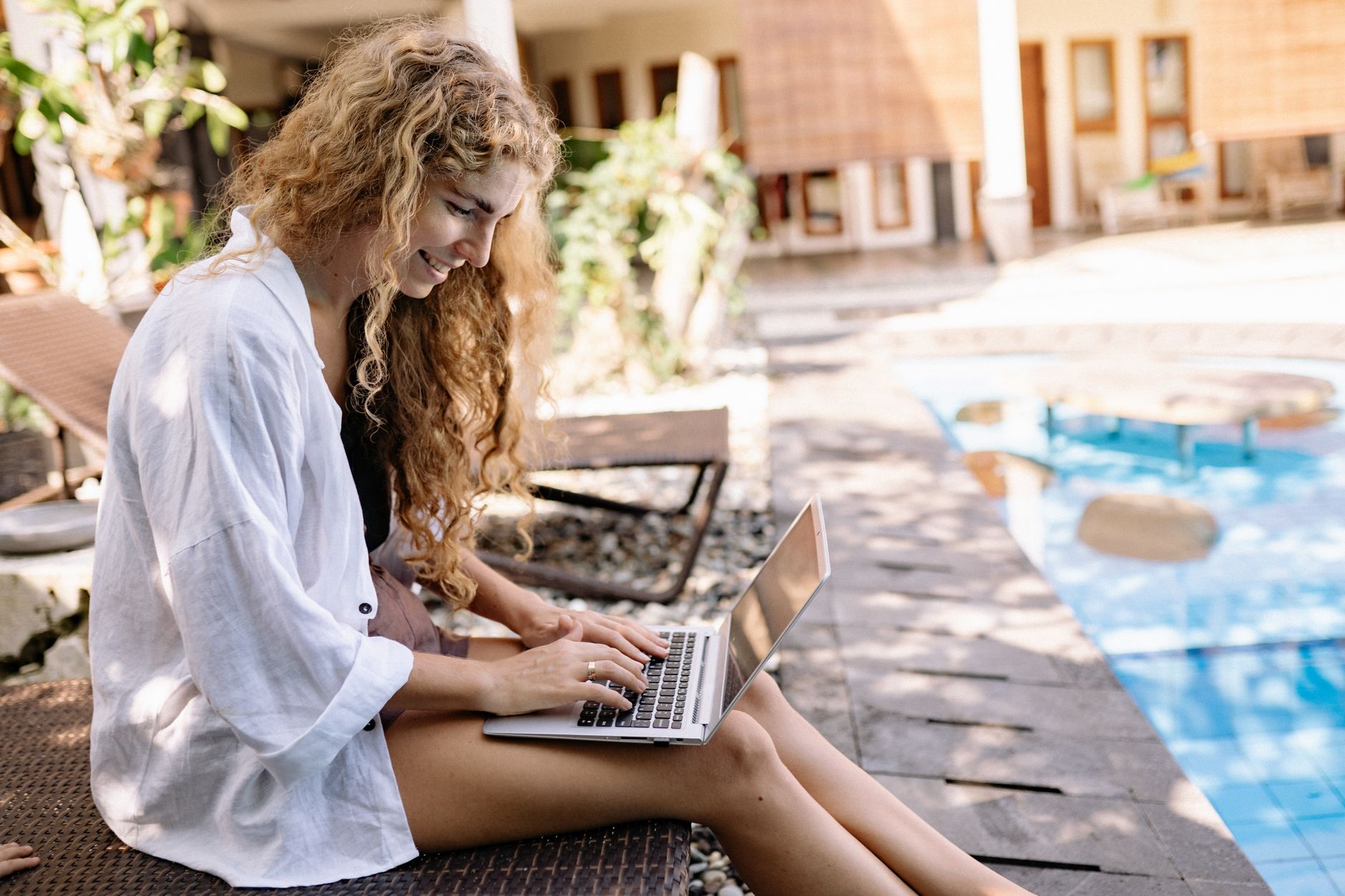 A smiling woman using a laptop while sitting by a poolside, enjoying a sunny day.