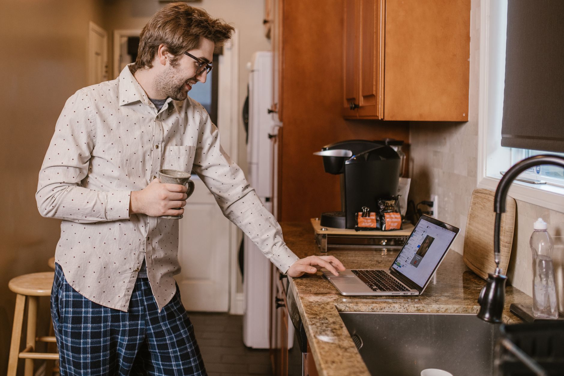 Man in pajamas using a laptop in kitchen, enjoying remote learning with a coffee.