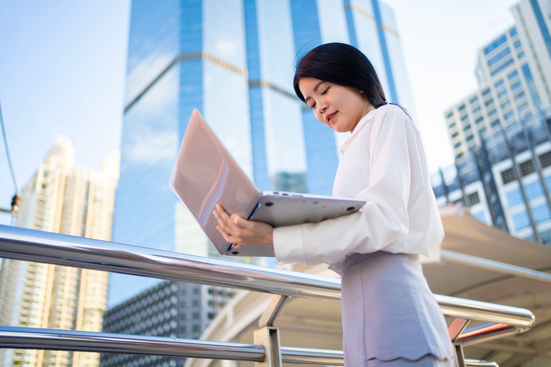 Asian businesswoman using laptop outdoors in Bangkok's modern cityscape.