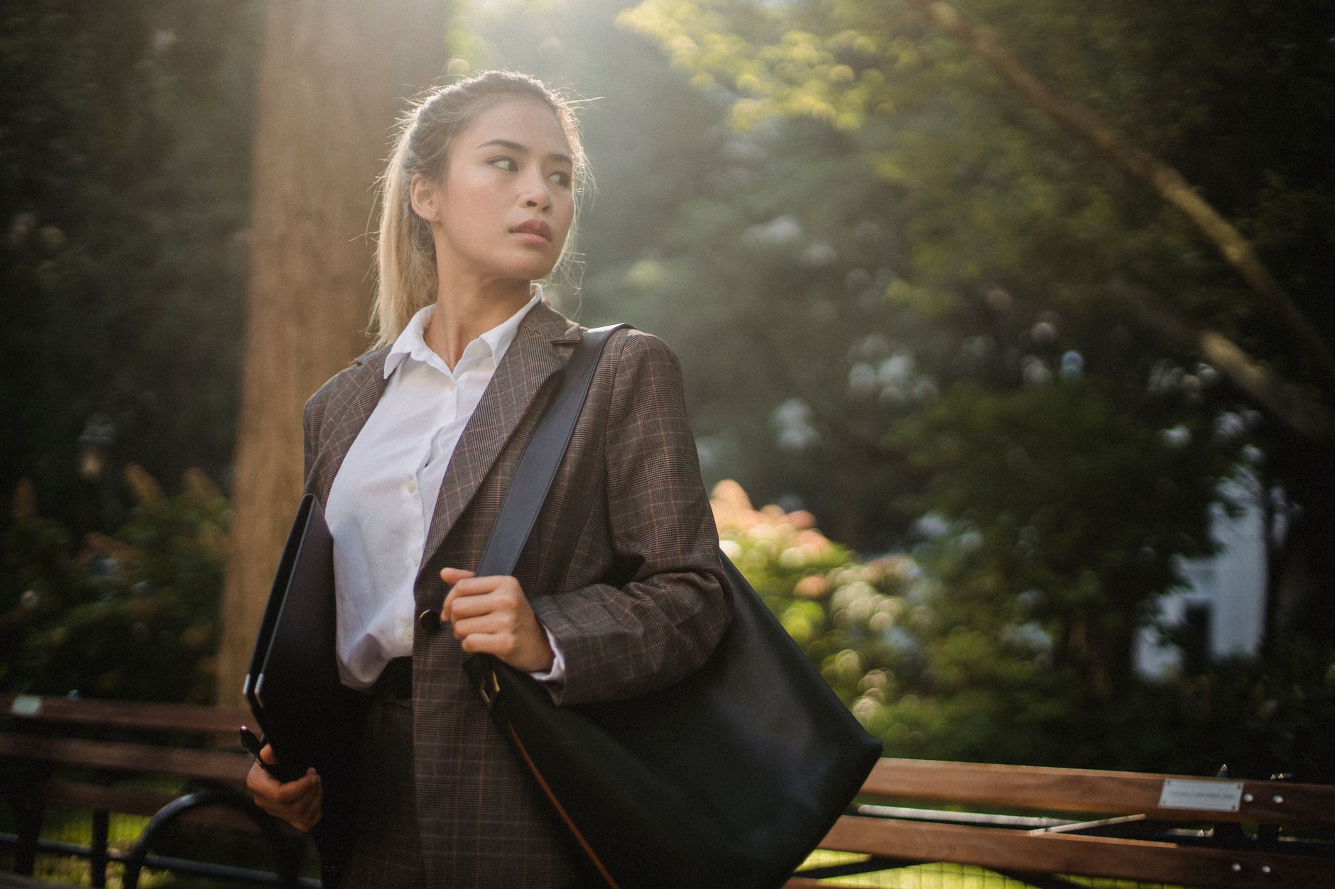 A professional woman in a suit stands confidently in a sunlit park, holding a laptop bag.