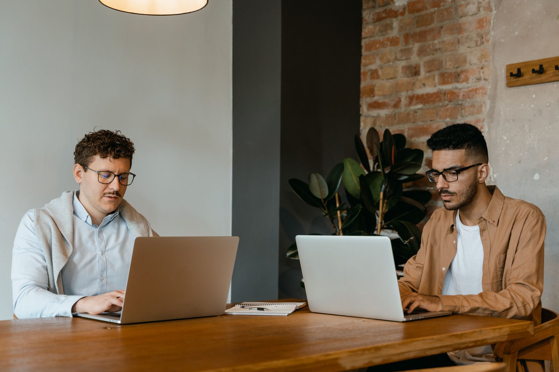 Two colleagues working on laptops in a modern office setting with a wooden table and brick wall.