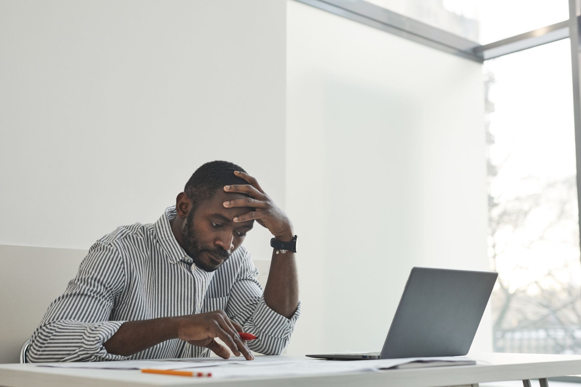 A thoughtful man sitting at a desk with a laptop and papers, contemplating work in a bright, modern office.