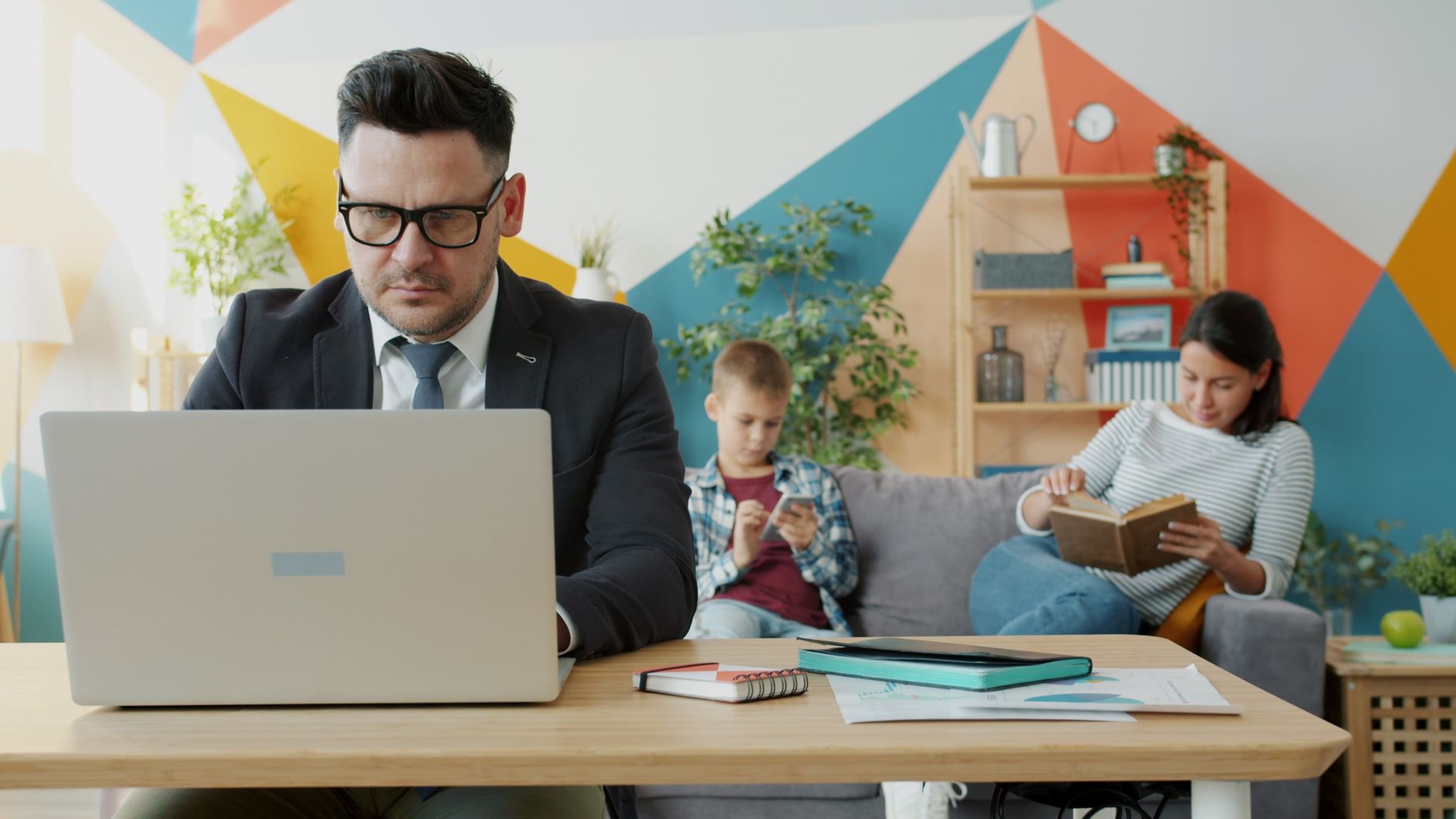 A man working on a laptop at home with family relaxing in the background.