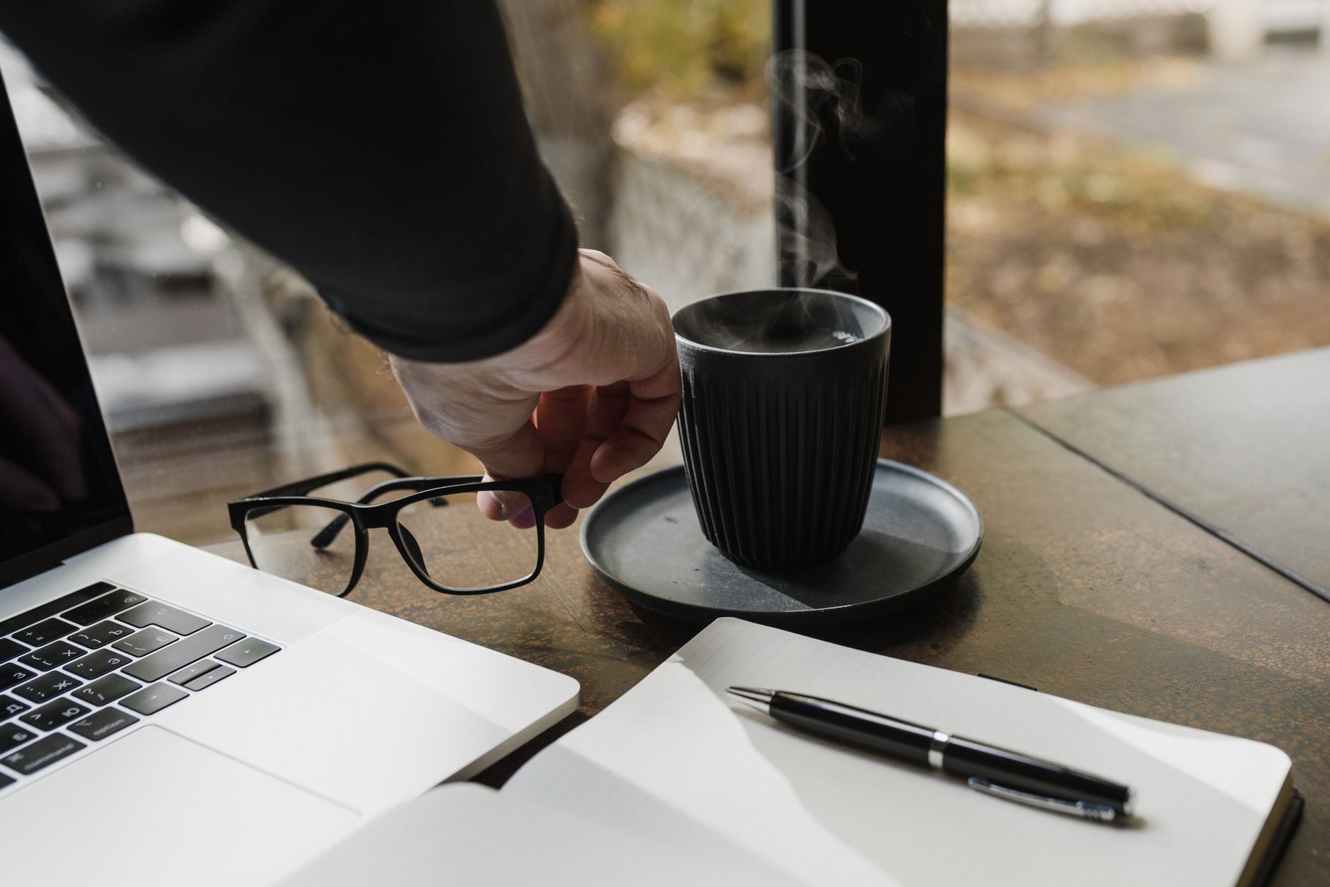 A hand reaches for a steaming cup of coffee near a laptop and open notebook, creating a cozy workspace vibe.