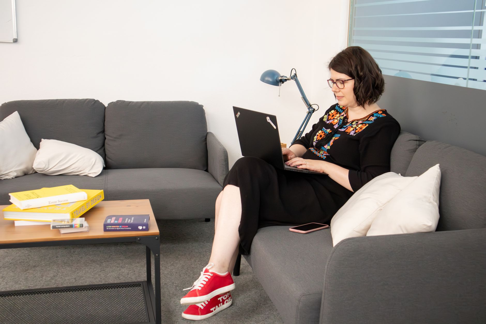 A woman sits on a couch working on her laptop in a cozy home office setup, surrounded by books and a lamp.