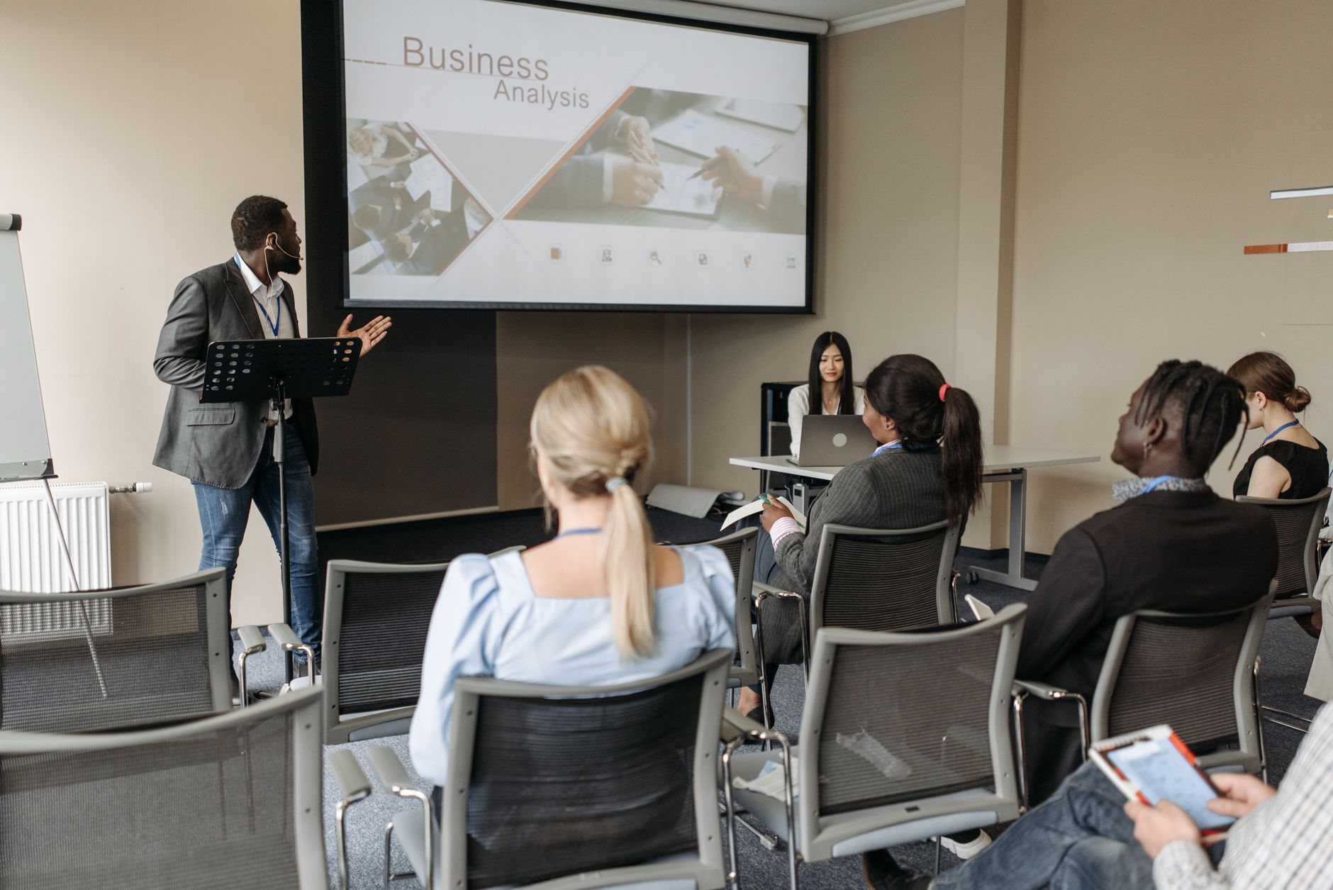 A diverse group attends a business analysis seminar with a presenter and projection screen in a modern conference room.