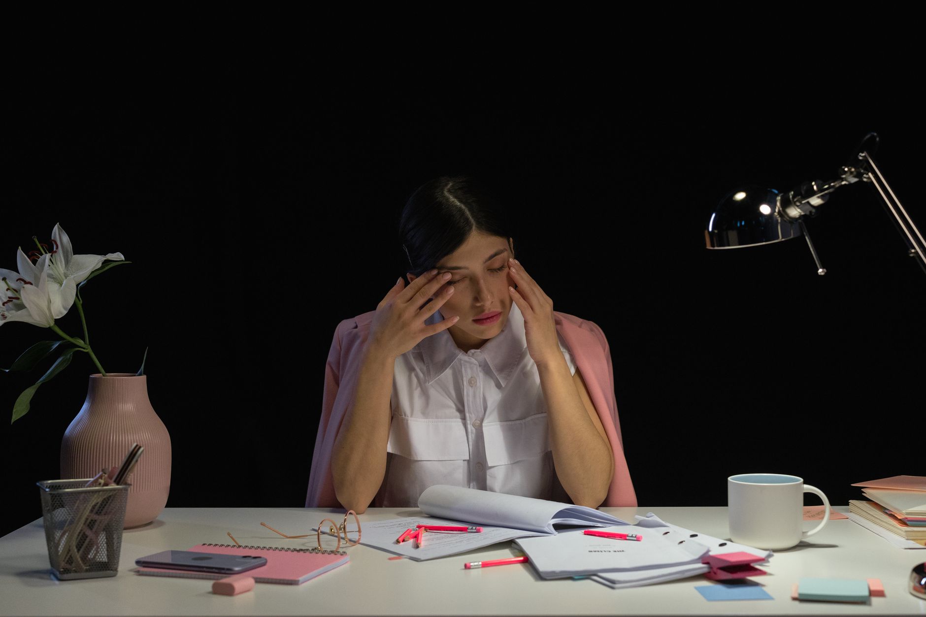 A tired woman sits at her office desk late at night, surrounded by papers and stationery.